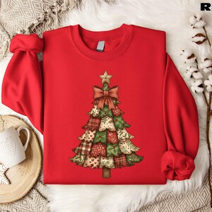A red sweater with a Christmas tree design printed on it, placed on a white fur rug. There is also a white mug and a branch of cotton balls nearby.