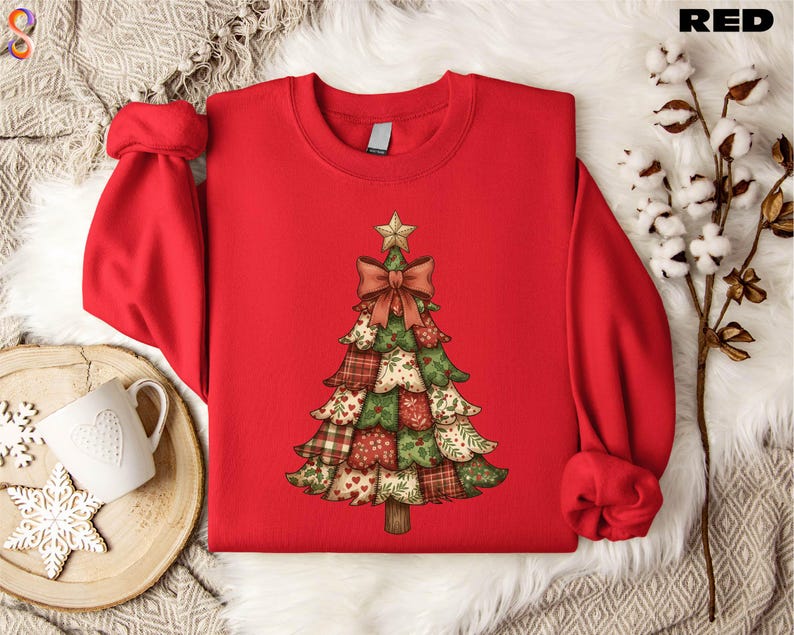 A red sweater with a Christmas tree design printed on it, placed on a white fur rug. There is also a white mug and a branch of cotton balls nearby.