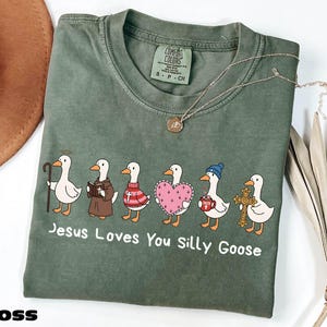 A green t-shirt with a graphic design of six cartoon ducks, each with a different outfit and accessory, along with the text "Jesus Loves You Silly Goose".