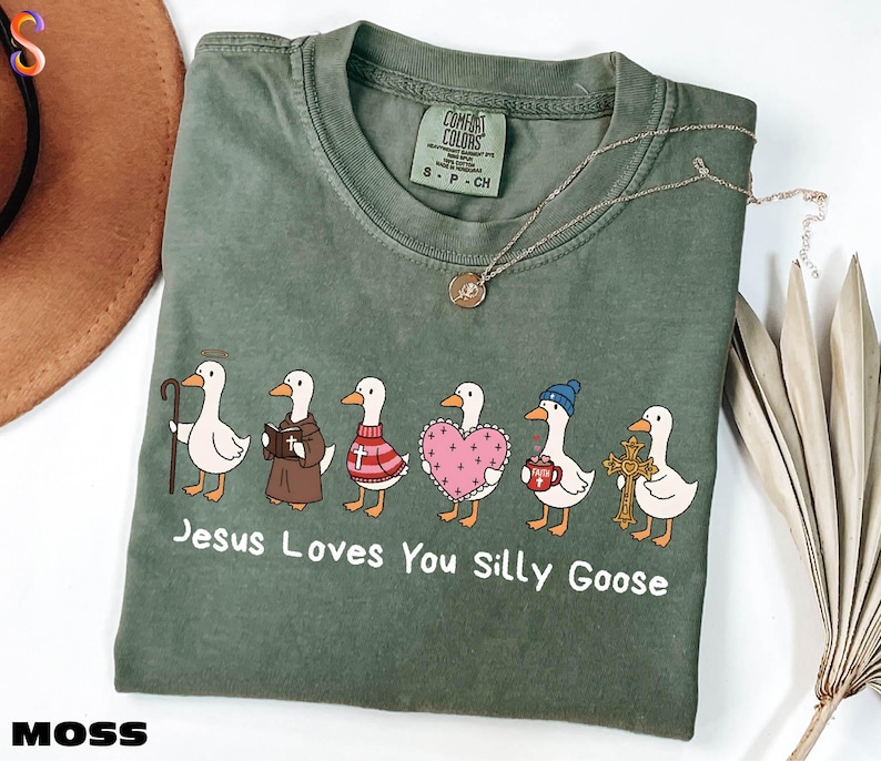 A green t-shirt with a graphic design of six cartoon ducks, each with a different outfit and accessory, along with the text "Jesus Loves You Silly Goose".