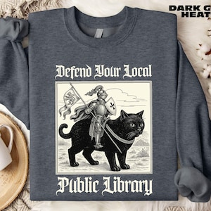 May include: Dark grey heather sweatshirt with a graphic of a knight riding a black cat, holding a flag and shield. The text reads "Defend Your Local Public Library". The sweatshirt has a crew neck and long sleeves.