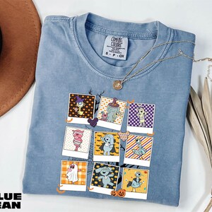A blue denim t-shirt with a graphic design of various cartoon characters, including a cat, dog, and other whimsical figures, is displayed on a white surface alongside a brown hat and some dried leaves.