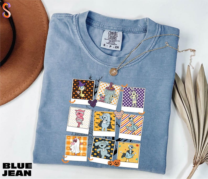 A blue denim t-shirt with a graphic design of various cartoon characters, including a cat, dog, and other whimsical figures, is displayed on a white surface alongside a brown hat and some dried leaves.