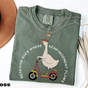 May include: Moss green t-shirt with a graphic of a goose riding a scooter, wearing a red cap. The text "BEHAVIOR GOES WHERE REINFORCEMENT FLOWS" is printed around the goose. A gold necklace is draped on the shirt.