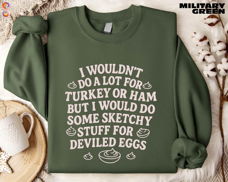 A green sweatshirt with white text printed on it, placed on a surface with a mug and some white flowers.