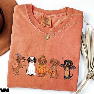 A pink t-shirt with a Halloween-themed graphic of five dogs dressed in costumes, including a witch, pumpkin, bat, and dog with a pumpkin.