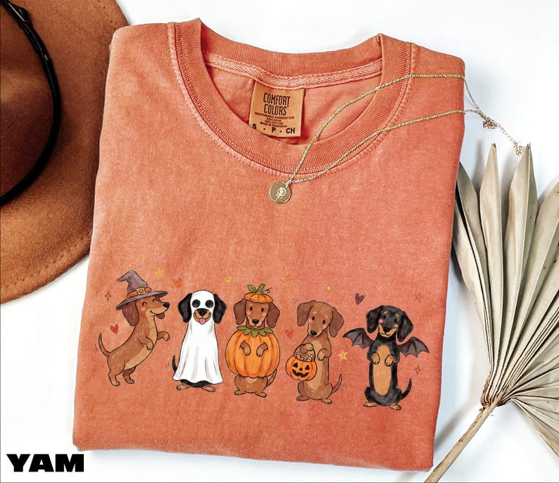 A pink t-shirt with a Halloween-themed graphic of five dogs dressed in costumes, including a witch, pumpkin, bat, and dog with a pumpkin.