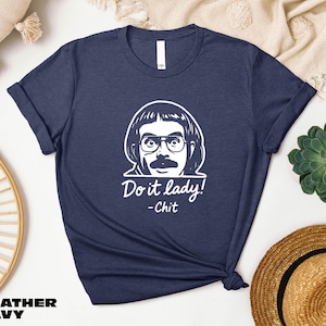 May include: Heather navy blue t-shirt with a white graphic of a man's face, mustache, and glasses. The text "Do it lady! - Chit" is below the graphic. The shirt is folded at the bottom.