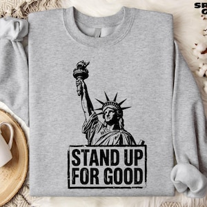 May include: Sport grey sweatshirt featuring a black and white graphic of the Statue of Liberty holding a torch. Below the statue is the text "STAND UP FOR GOOD" in a black, rectangular box. The sweatshirt has a crew neck and long sleeves.