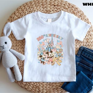 May include: White t-shirt featuring a colorful graphic celebrating the "250th Anniversary" with Mickey Mouse, Minnie Mouse, Donald Duck, and Goofy. The shirt is paired with blue denim jeans and a gray stuffed bunny.