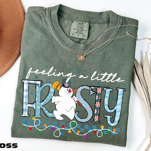 A green t-shirt with a snowman graphic and the text "feeling a little FROSTY" printed on it, along with a necklace and a hat.