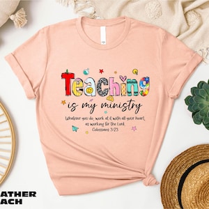 May include: Heather peach-colored t-shirt with the text "Teaching is my ministry" in colorful, playful lettering. The shirt also features the quote "Whatever you do, work at it with all your heart, as working for the Lord, Colossians 3:23."