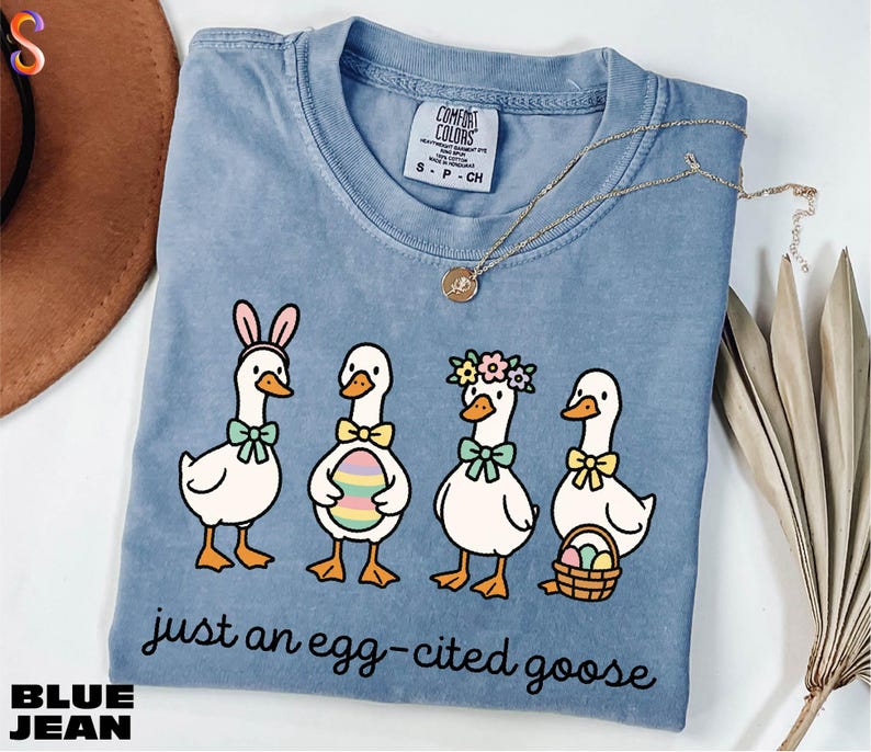 A blue t-shirt with a cartoon illustration of four cartoon ducks wearing Easter-themed accessories, accompanied by the text "Just an egg-cited goose".