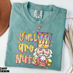 May include: Light green t-shirt with the text "Y'all are Nuts" and a colorful nutcracker graphic. The shirt has a crew neck and short sleeves. A silver necklace with a pendant is draped on the shirt.