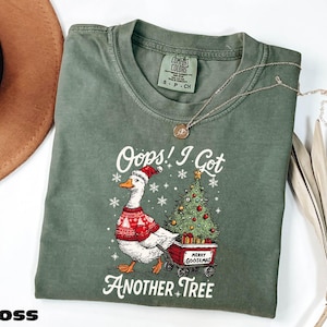 Puede incluir: Camiseta verde musgo con un gráfico de un ganso con un suéter y gorro navideños, tirando de un carro con un árbol de Navidad. El texto dice "Oops! I Got Another Tree" y "Merry Goosemas".