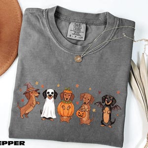 A gray t-shirt with a Halloween-themed graphic of five dachshund dogs dressed in costumes, including a witch, pumpkin, bat, and pumpkin, along with a necklace and a brown hat.