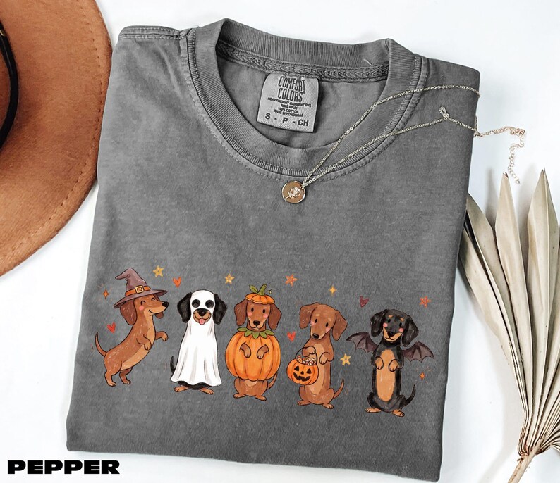 A gray t-shirt with a Halloween-themed graphic of five dachshund dogs dressed in costumes, including a witch, pumpkin, bat, and pumpkin, along with a necklace and a brown hat.