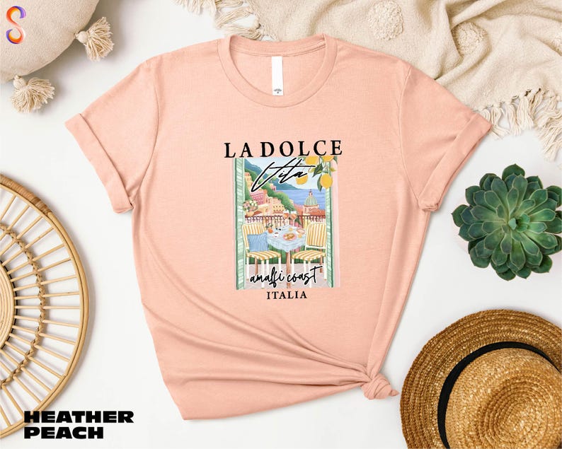a pink t-shirt with a graphic design of a coastal scene, along with the text "LADOLCE VITA ANTIGIO ITALIA". The t-shirt is displayed on a white background, surrounded by various accessories such as a straw hat, a woven basket, and a potted plant.