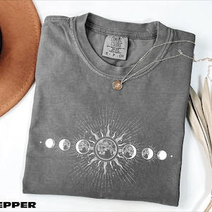 May include: A gray Comfort Colors t-shirt featuring a white graphic of the sun and moon phases. The shirt has a crew neck and short sleeves. A silver necklace with a round pendant rests on the shirt. A brown hat is in the upper left corner.