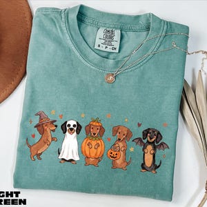 A light green t-shirt with a Halloween-themed graphic of five dogs dressed in costumes, including a witch, pumpkin, bat, and dog with a pumpkin.
