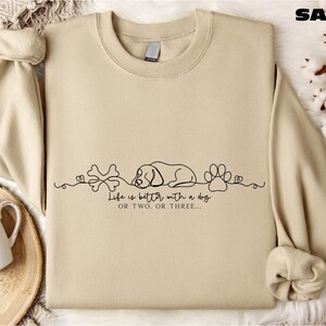 a beige sweatshirt with a graphic design on it, including a sleeping dog, bones, and paw prints, along with the text "Life is better with a dog" or "Life is better with two or three".