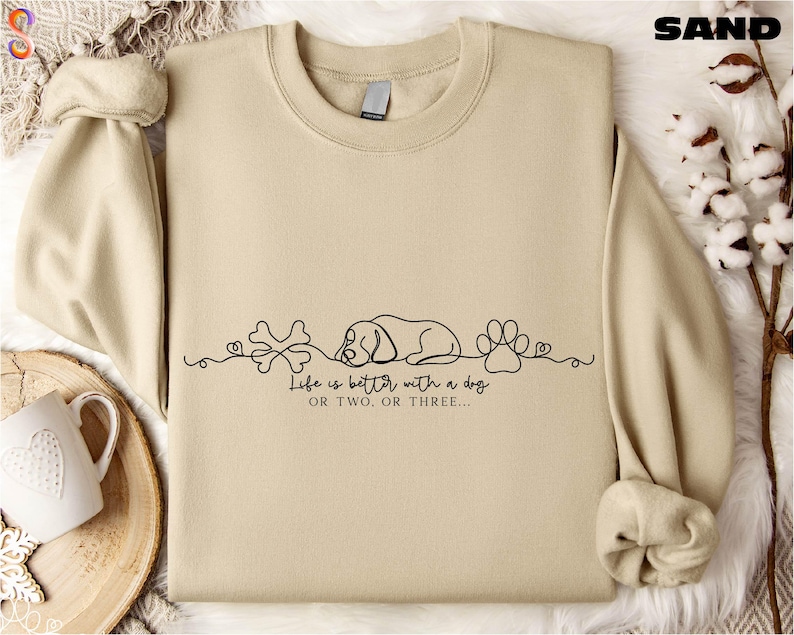 a beige sweatshirt with a graphic design on it, including a sleeping dog, bones, and paw prints, along with the text "Life is better with a dog" or "Life is better with two or three".