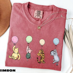 A red t-shirt with a design of five cartoon characters from Winnie the Pooh, including Piglet, Tigger, Eeyore, and Tigger, each holding a balloon. The t-shirt is placed on a white surface, with a brown hat and some dried leaves nearby.