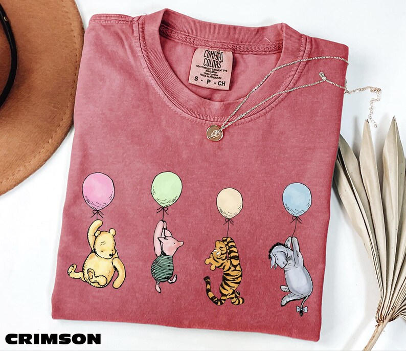 A red t-shirt with a design of five cartoon characters from Winnie the Pooh, including Piglet, Tigger, Eeyore, and Tigger, each holding a balloon. The t-shirt is placed on a white surface, with a brown hat and some dried leaves nearby.