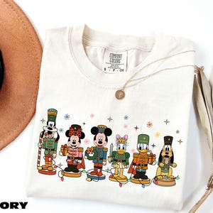 A white t-shirt with a graphic design of Disney characters, including Mickey Mouse, Minnie Mouse, Donald Duck, Goofy, and Pluto, along with a necklace and a hat.
