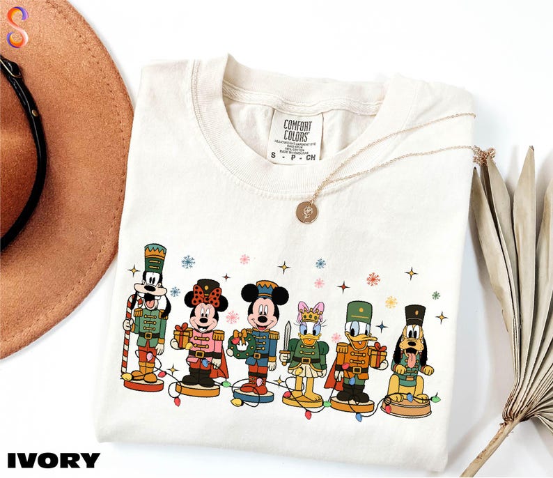 A white t-shirt with a graphic design of Disney characters, including Mickey Mouse, Minnie Mouse, Donald Duck, Goofy, and Pluto, along with a necklace and a hat.