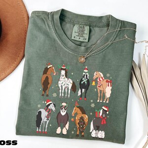 A green t-shirt with a graphic of nine horses wearing Christmas-themed outfits, including Santa hats and scarves, is displayed on a white surface alongside a brown hat and some dried leaves.