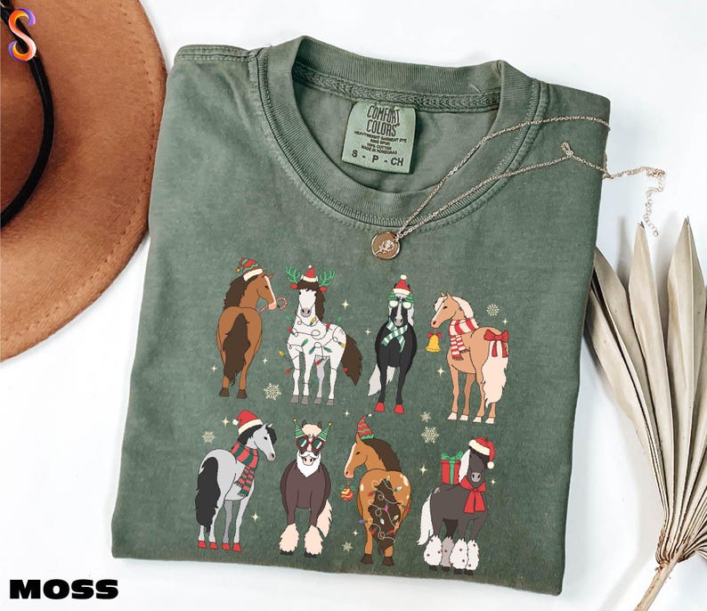 A green t-shirt with a graphic of nine horses wearing Christmas-themed outfits, including Santa hats and scarves, is displayed on a white surface alongside a brown hat and some dried leaves.