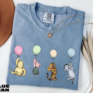 A blue t-shirt with illustrations of characters from the Winnie the Pooh series, including Piglet, Tigger, and Eeyore, along with balloons. The t-shirt is placed on a white surface, with a brown hat and some leaves nearby.