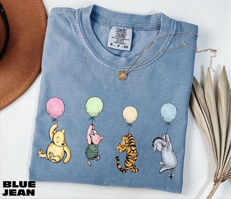 A blue t-shirt with illustrations of characters from the Winnie the Pooh series, including Piglet, Tigger, and Eeyore, along with balloons. The t-shirt is placed on a white surface, with a brown hat and some leaves nearby.