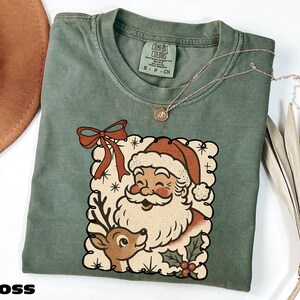 A green t-shirt with a Santa Claus graphic and a bow, along with a necklace and a hat, are displayed on a white surface.