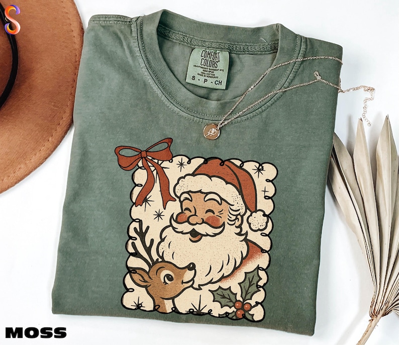 A green t-shirt with a Santa Claus graphic and a bow, along with a necklace and a hat, are displayed on a white surface.