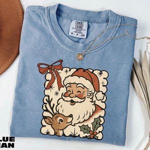 A blue t-shirt with a Santa Claus and reindeer graphic, along with a necklace and a hat, are displayed on a white surface.