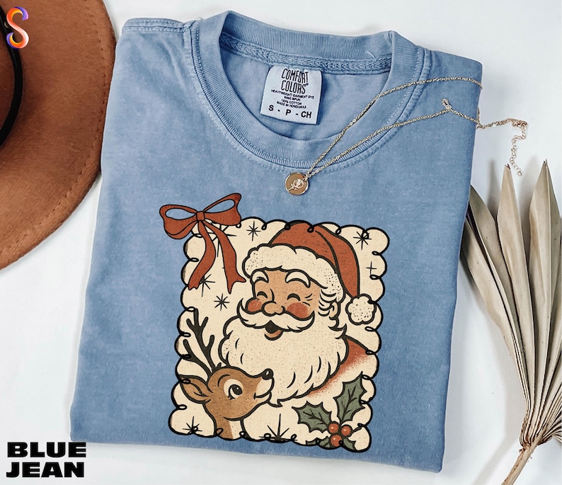 A blue t-shirt with a Santa Claus and reindeer graphic, along with a necklace and a hat, are displayed on a white surface.
