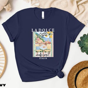 a navy blue t-shirt with a graphic design of a coastal town, along with the text "LADOICE ITALIA" printed on it. The t-shirt is displayed on a white background, surrounded by various objects such as a straw hat, a woven basket, and a potted plant.