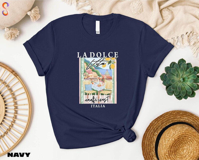 a navy blue t-shirt with a graphic design of a coastal town, along with the text "LADOICE ITALIA" printed on it. The t-shirt is displayed on a white background, surrounded by various objects such as a straw hat, a woven basket, and a potted plant.