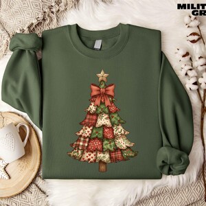 A green sweater with a Christmas tree design printed on it, placed on a white fur rug. There is also a mug and some white flowers in the background.
