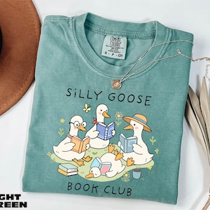 May include: Light green t-shirt with the text "SILLY GOOSE BOOK CLUB" and a graphic of cartoon geese reading books. The geese are white with orange beaks and feet, and some wear glasses or hats. A small butterfly and flowers are also in the design.