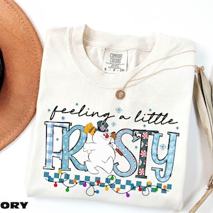 A white t-shirt with a snowman graphic and the text "feeling a little FROSTY" printed on it, along with a necklace and a hat.