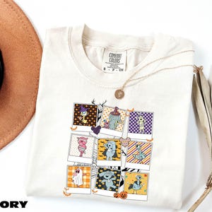 a white t-shirt with a graphic design of various cartoon characters, including a ghost, a skeleton, and a pumpkin. The t-shirt is placed on a white surface, with a brown hat and a fan visible in the background.