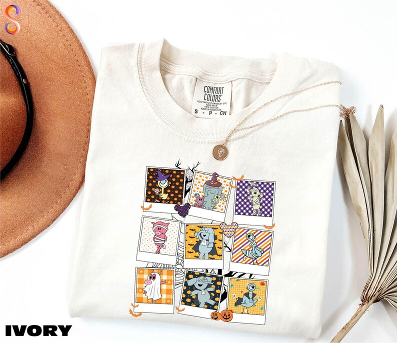 a white t-shirt with a graphic design of various cartoon characters, including a ghost, a skeleton, and a pumpkin. The t-shirt is placed on a white surface, with a brown hat and a fan visible in the background.