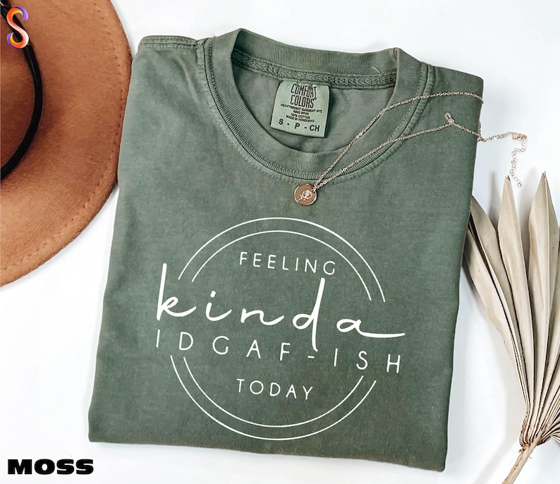 a green t-shirt with a white text design that says "Feeling kinda idgafish today" and a small charm on the neck. The t-shirt is laid out on a white surface, with a brown hat and some dried leaves nearby.