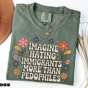 Op de afbeelding: Mosgroen t-shirt met de tekst "IMAGINE HATING IMMIGRANTS MORE THAN PEDOPHILES" in een retro lettertype, omringd door kleurrijke bloemen. Een gouden ketting ligt op het shirt. Het shirt is op een wit oppervlak gevouwen.