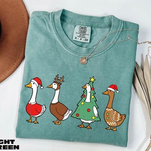 A light green t-shirt with a Christmas-themed graphic of four cartoon ducks dressed in festive outfits, including Santa hats and Christmas trees.