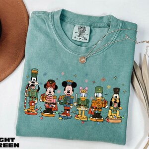 A light green t-shirt with a graphic of Disney characters, including Minnie Mouse, Mickey Mouse, Donald Duck, Goofy, and Pluto, standing on a green base. The t-shirt is displayed on a white background, along with a brown hat and some dried leaves.