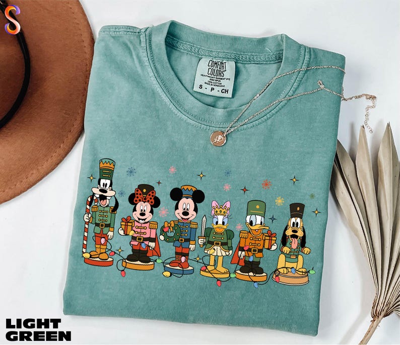 A light green t-shirt with a graphic of Disney characters, including Minnie Mouse, Mickey Mouse, Donald Duck, Goofy, and Pluto, standing on a green base. The t-shirt is displayed on a white background, along with a brown hat and some dried leaves.